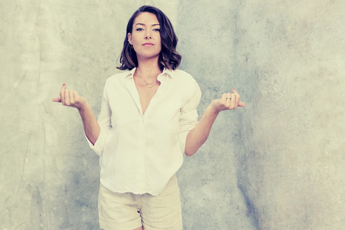 Ashlie Walker in a white shirt, shrugging with a smile against a grey wall
