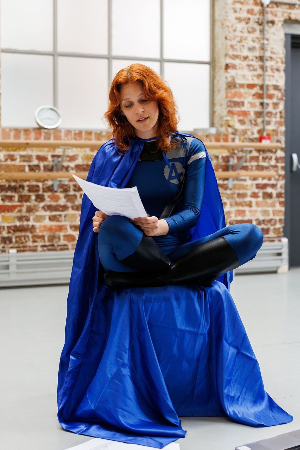 An actor in a blue cape reading a script in a brick-walled rehearsal room