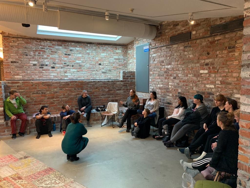 A group sitting in a circle on the floor of a brick-walled studio during a workshop